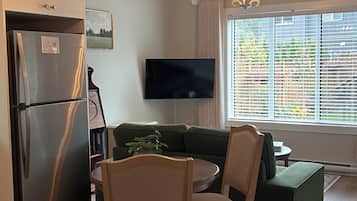 Een moderne woonkamer met een eethoek, een flatscreen televisie, een groot raam met zonwering en een inbouwkoelkast.