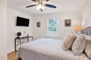 A bedroom with a bed, a ceiling fan, a TV, a window, and a bedside table with a lamp. - Oakleighs Little Cottage (Mobile)