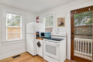 A compact kitchen with a refrigerator, microwave, and oven, a window with blinds, and a door with a view of a wooden fence. - Oakleighs Little Cottage (Mobile)