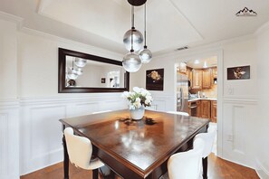 A dining area with a wooden table, white chairs, a chandelier, and a kitchen in the background. - Misty Mornings at Sugar Top (Sugar Mountain)