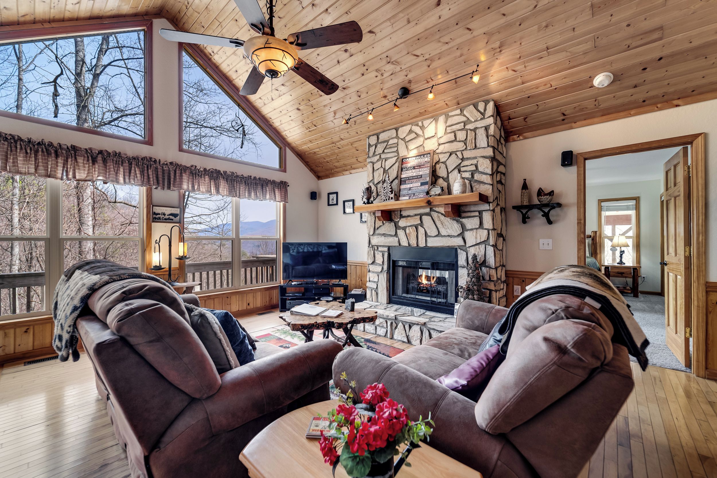 A cozy living room with a stone fireplace, comfortable seating, a flat-screen TV, and a large window overlooking a scenic view.