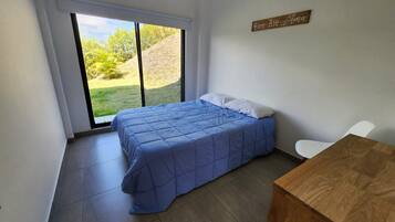 Een slaapkamer met een bed, een houten tafel, een witte stoel en uitzicht op een grasveld door een raam.