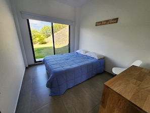 Un dormitorio con una cama, una mesa de madera, una silla blanca y vistas a un área verde por una ventana.