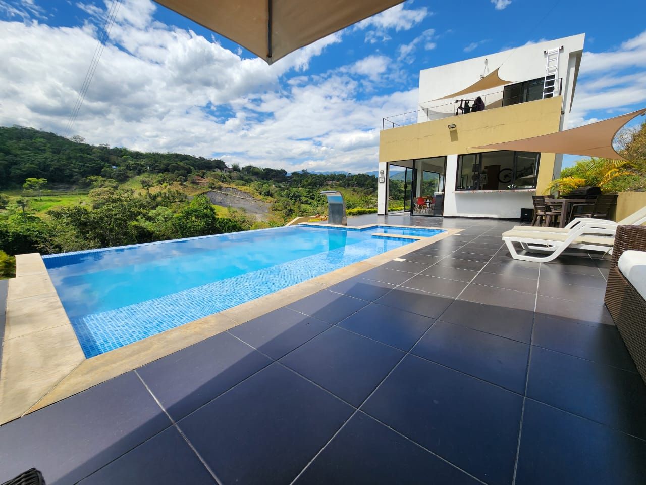 A modern outdoor swimming pool with a view of a lush landscape, a two-story building with large windows, and a covered seating area.