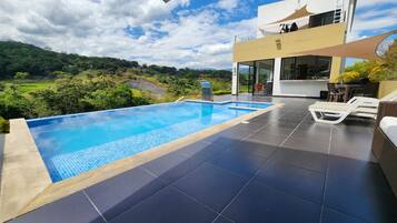 A modern outdoor swimming pool with a view of a lush landscape, a two-story building with large windows, and a covered seating area.