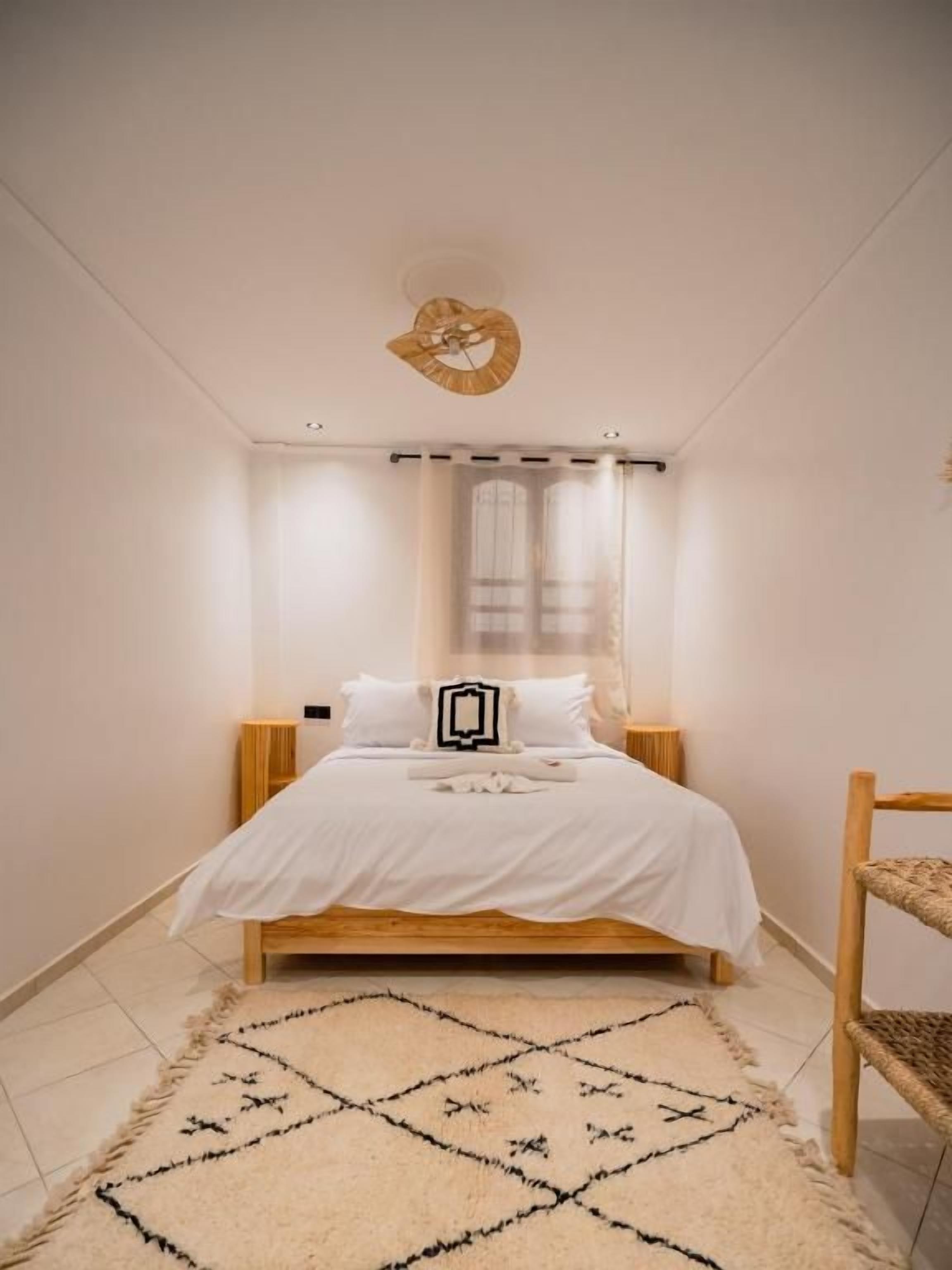 A bedroom with a bed, a headboard, a bedside table, a chair, a window with curtains, and a patterned rug.