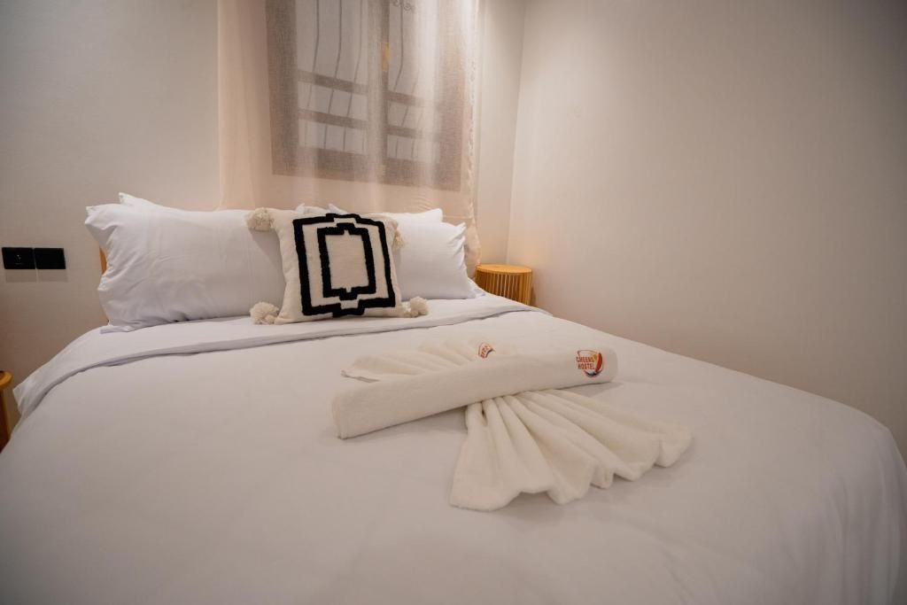 Una cama bien hecha con ropa blanca y una toalla, un almohadón con diseño blanco y negro, y una ventana con cortinas traslúcidas.