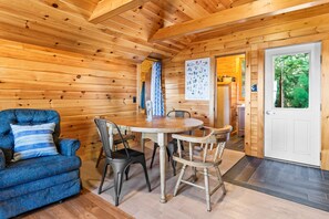 A wooden cabin interior with a dining table, chairs, a sofa, and a door leading outside. - The Cabins on Webb Pond - NEW! Waterfront (Eastbrook)