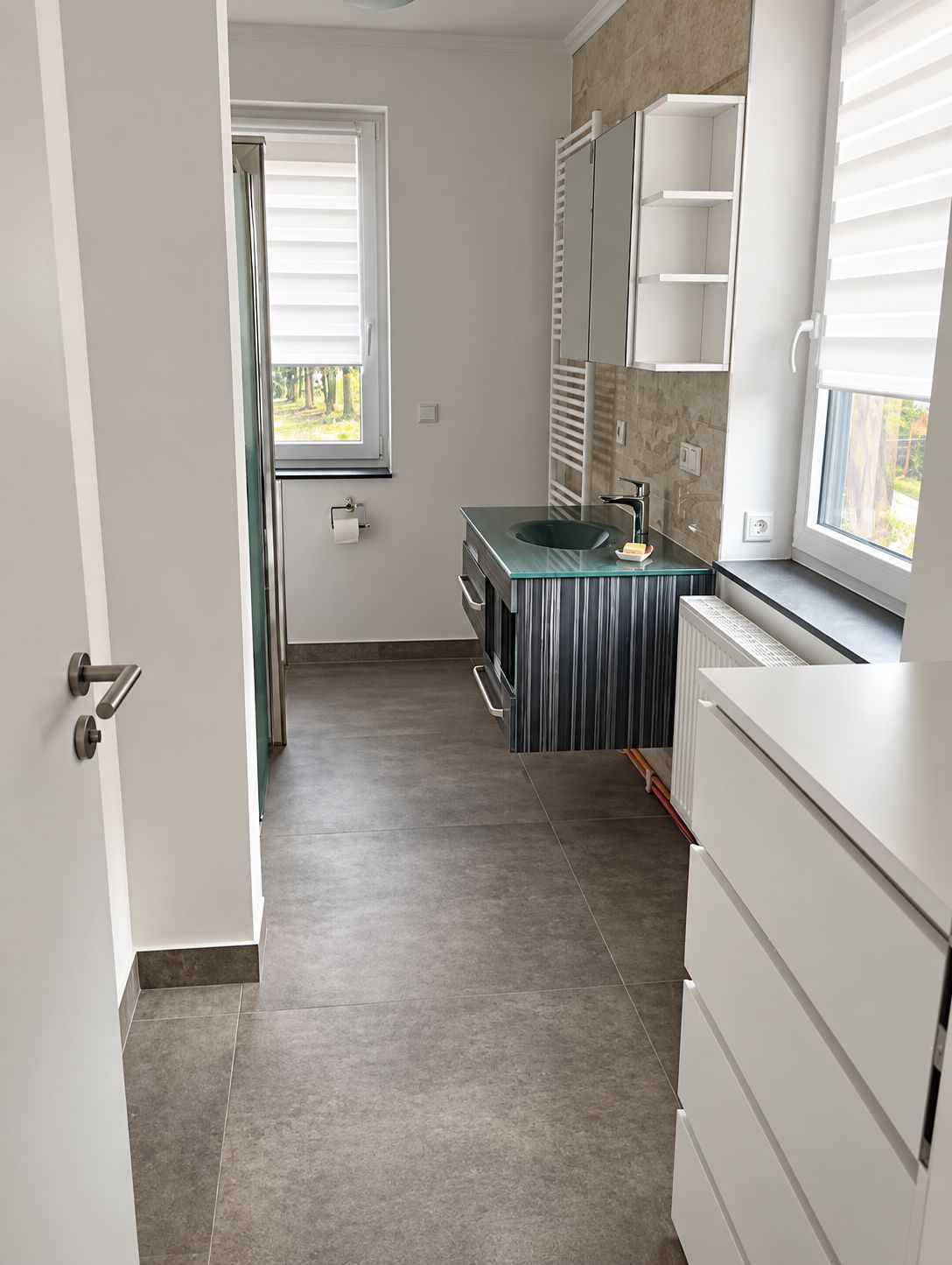 A modern bathroom with a glass sink, white cabinetry, and a window with blinds.