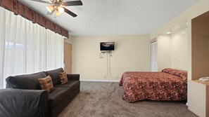 Habitación de hotel con un sofá marrón, cama con colcha estampada, ventilador de techo, televisor montado y piso alfombrado.