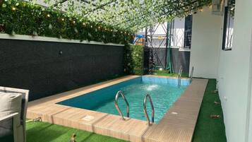 Una piscina con muelle de madera, rodeada de césped artificial y una pared negra.