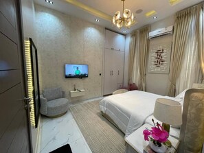 A hotel room with a bed, a television, a chair, a desk, and a decorative wall piece. - Luxury 5 BR Villa With Cinema and Pool (Lekki)