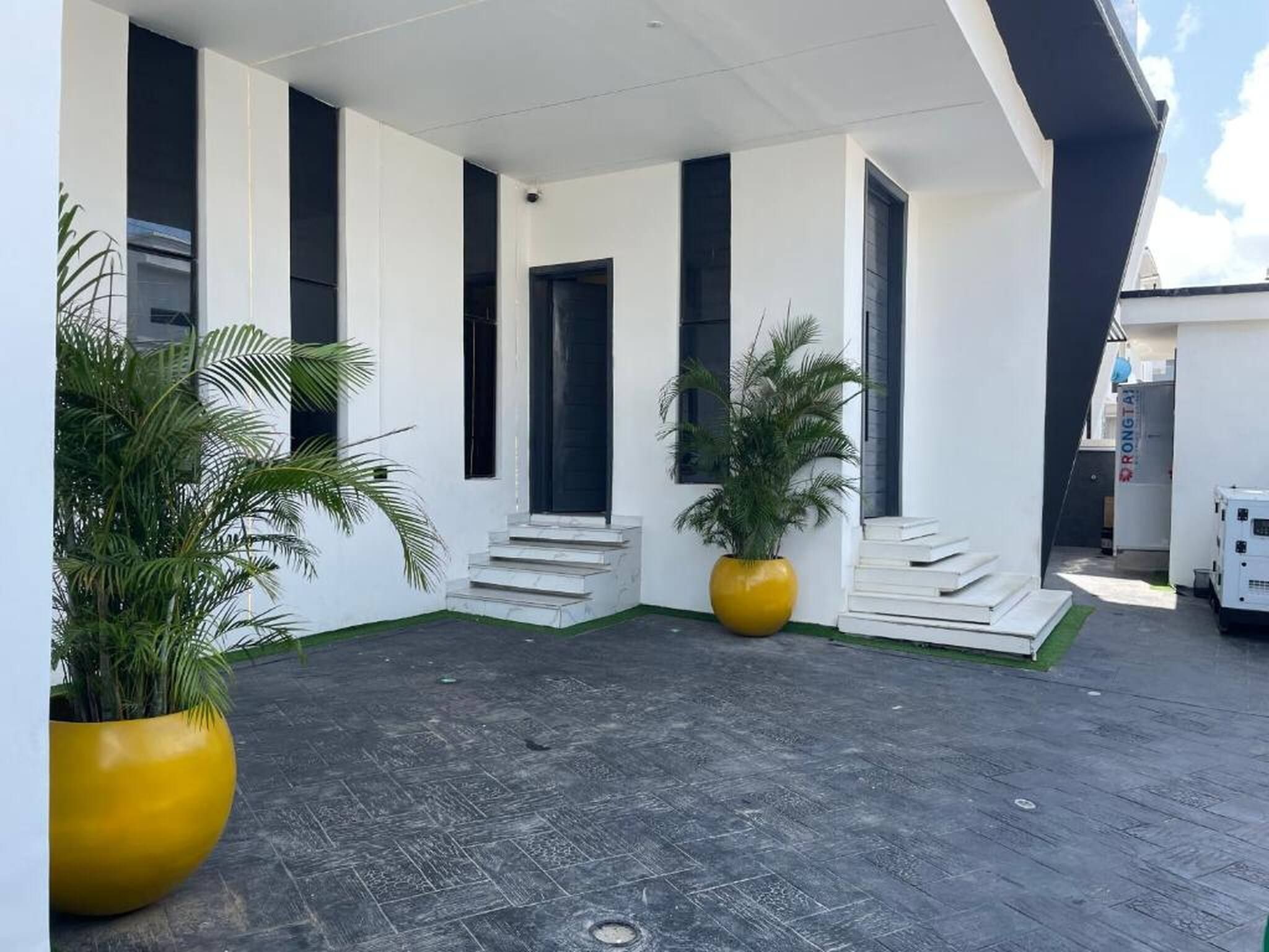 Un edificio moderno con fachada blanca, marcos de ventana negros y suelo de baldosas grises. Hay dos macetas grandes amarillas con plantas verdes.