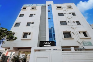 Edifício com vários andares, um elevador de vidro central, diversas janelas com grades de metal e uma placa indicando os serviços 'Collection' e 'Oyo-Serviced'.
