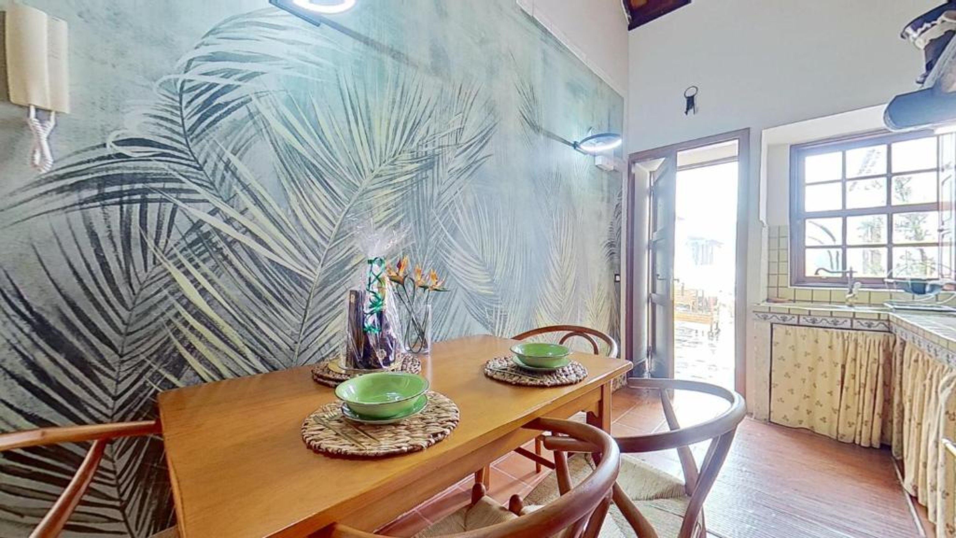 Un coin repas avec une table en bois, deux bols verts et des chaises. Un grand motif représentant des feuilles orne le mur derrière la table.