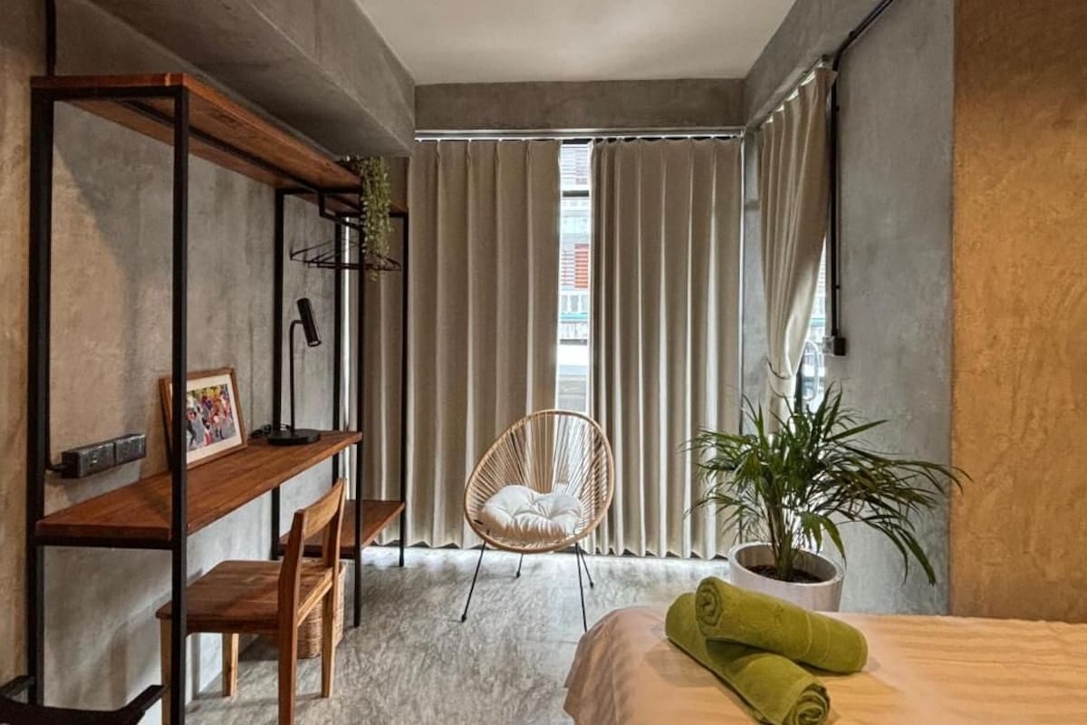 Ein modernes Schlafzimmer mit einem Bett, einer Holzbank, einem Korbsessel, einer Topfpflanze und einem großen Fenster mit Vorhängen.