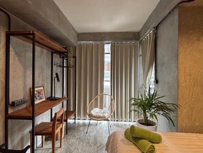 Ein modernes Schlafzimmer mit einem Bett, einer Holzbank, einem Korbsessel, einer Topfpflanze und einem großen Fenster mit Vorhängen.