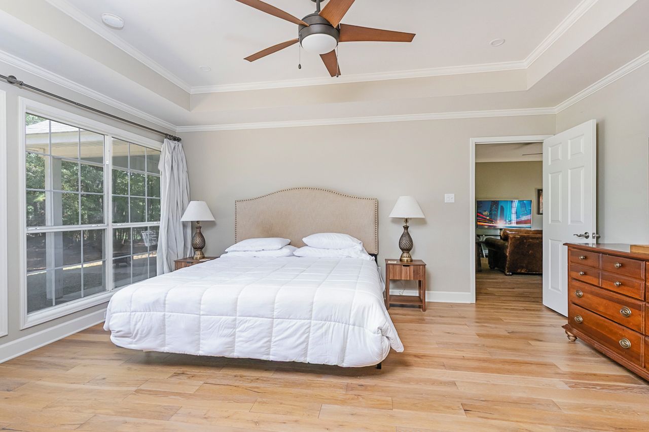 A bedroom with a large bed, two bedside lamps, a ceiling fan, and a view through a large window.
