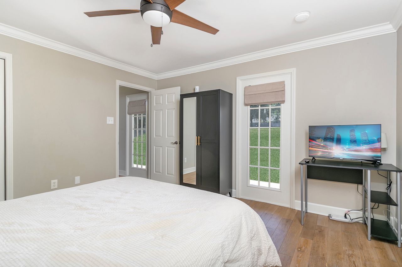 Une chambre à coucher avec un lit, un ventilateur de plafond, un téléviseur posé sur un meuble et une porte donnant sur un balcon.