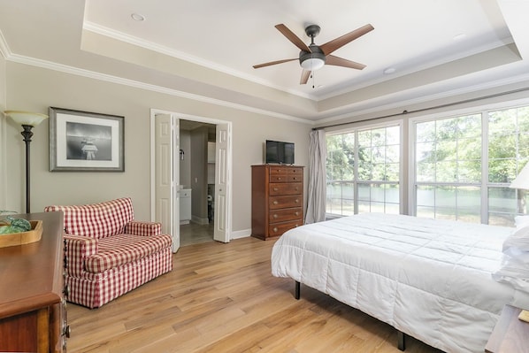 Un dormitorio con cama, cómoda, sofá rojo y blanco a cuadros, ventilador de techo y ventana con vistas a la vegetación.