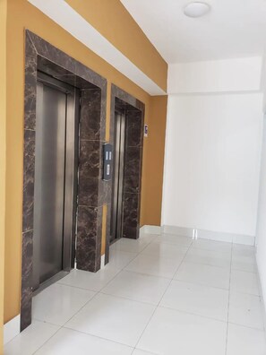 Un pasillo con dos ascensores, suelo de baldosas blancas y una pared de acento amarilla.