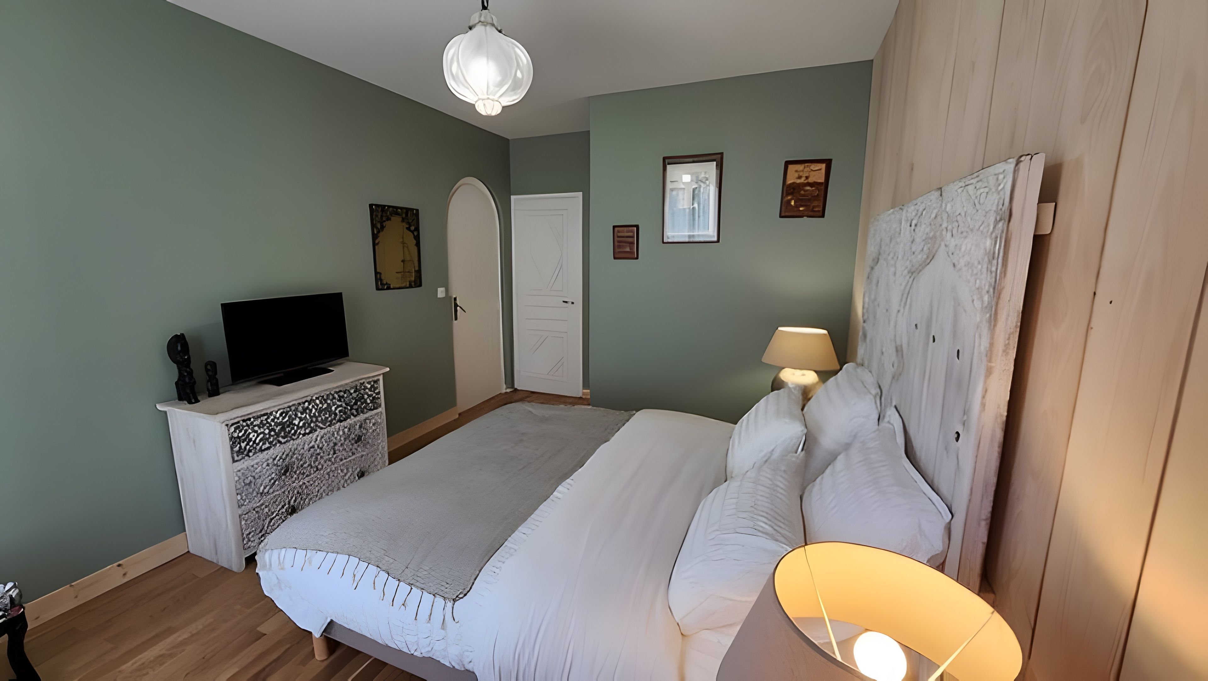 Une chambre à coucher avec un lit, un téléviseur posé sur une commode, une porte et un mur orné de tableaux.