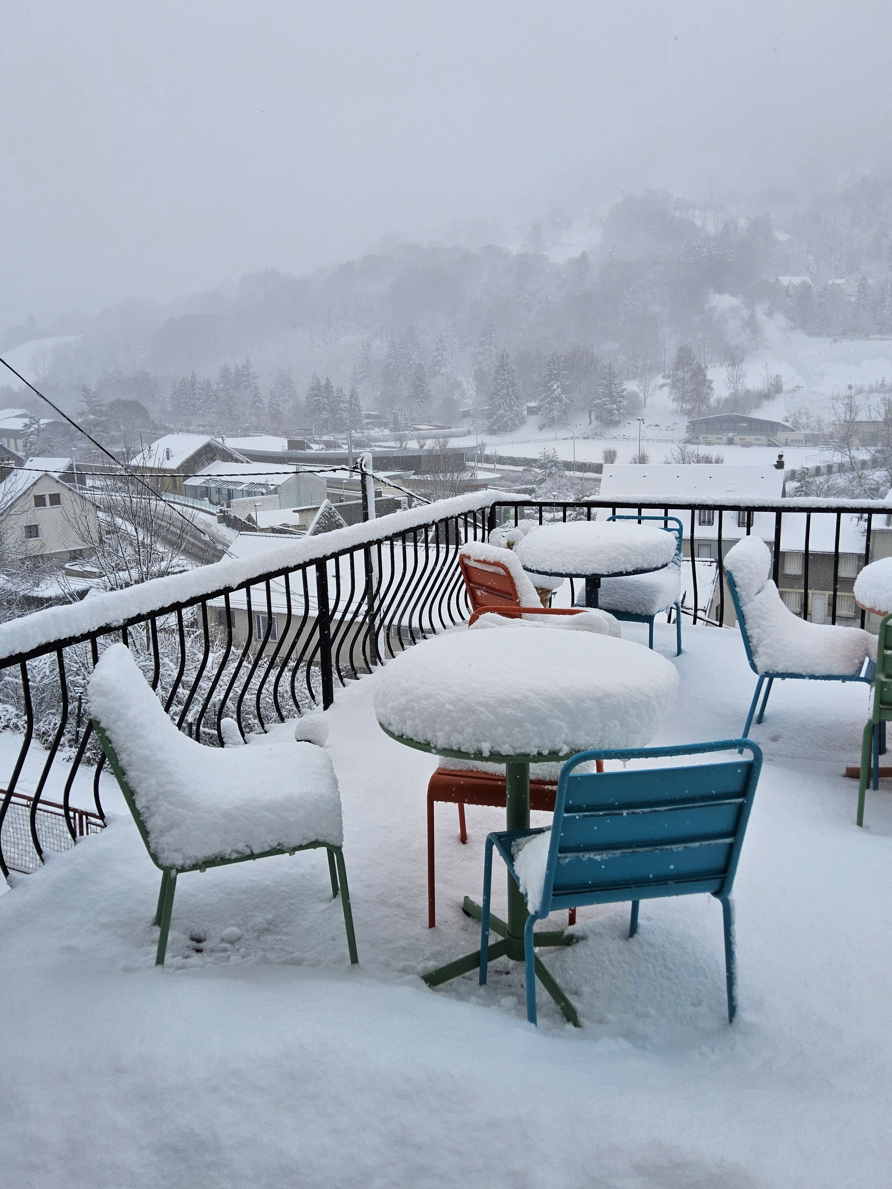 Buiten zitplaatsen met sneeuw bedekte tafels en stoelen, een metalen hekwerk en een uitzicht op een besneeuwd dorp.