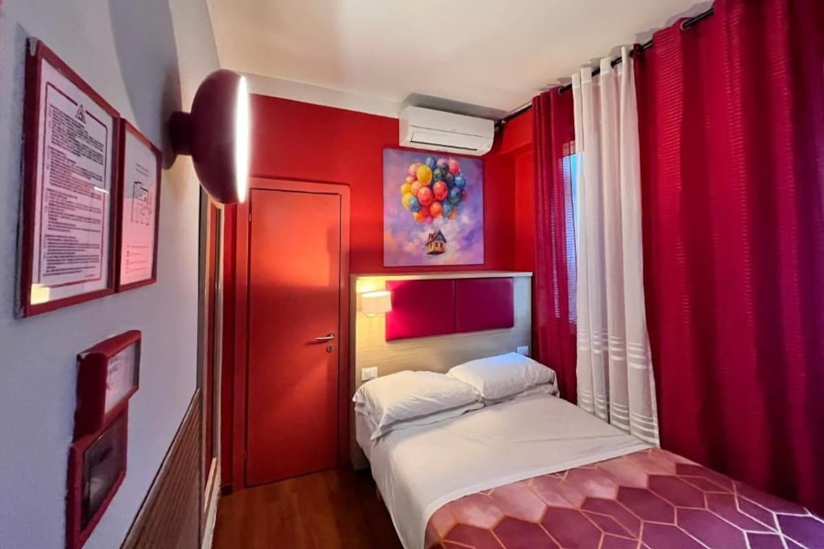 A hotel room with a bed, a painting, red curtains, and an air conditioning unit.