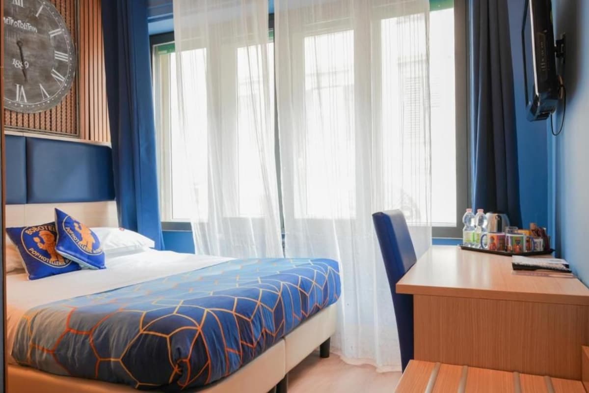 A hotel room with a bed, a desk, a chair, a clock, and a window with curtains.