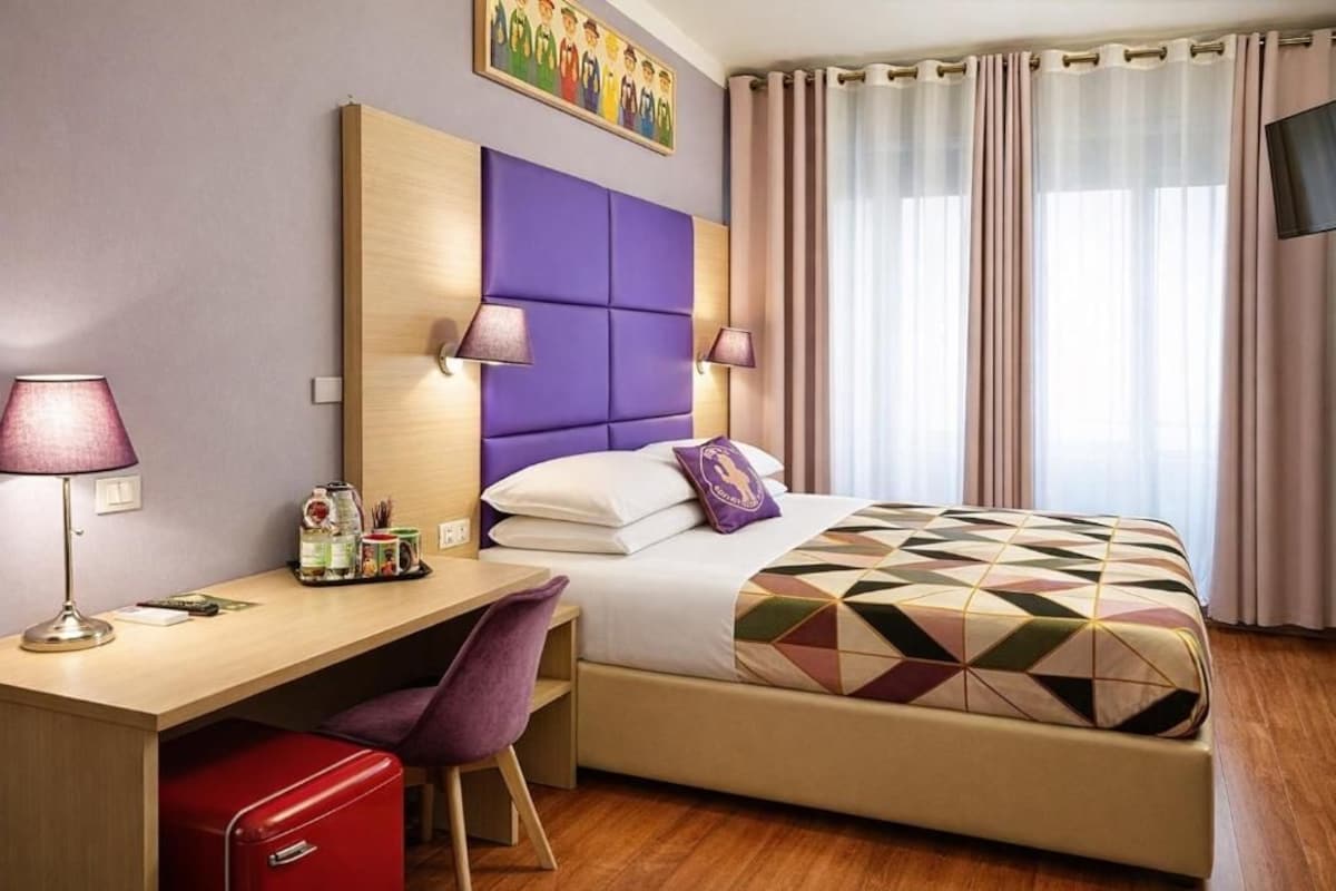 A hotel room with a bed, a desk with a chair, a TV, and a window with curtains.