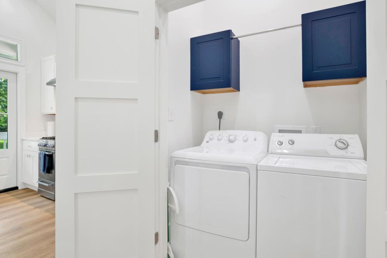 A laundry room with a washing machine and dryer, a white door, and a view into the kitchen area.