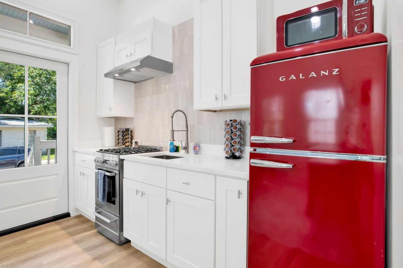 Una cocina moderna con armarios blancos, un refrigerador Galanz rojo, estufa de acero inoxidable y una ventana con vista a la vegetación.