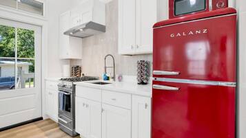 Una cocina moderna con armarios blancos, un refrigerador Galanz rojo, estufa de acero inoxidable y una ventana con vista a la vegetación.