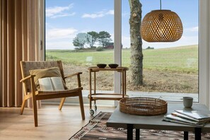 Ein Zimmer mit einem großen Fenster, einem Korbstuhl, einem kleinen Tisch mit einer Schale und einer hängenden Lampe.