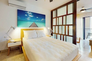 A hotel room with a bed, a nightstand, a desk lamp, a painting of a beach, and a large window with a view. - 1BR Condo Rooftop Pool & Gym Playa (Playa del Carmen)