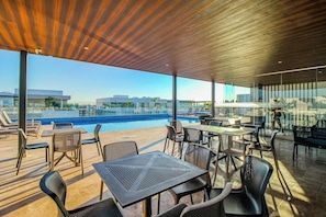 A rooftop bar with tables and chairs, overlooking a swimming pool and cityscape. - 1BR Condo Rooftop Pool & Gym Playa (Playa del Carmen)