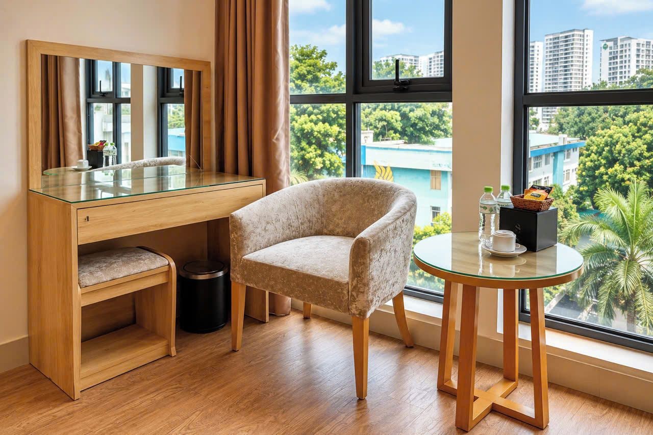 A room with a wooden desk, a cushioned chair, a small round table with a glass top, and a large window offering a view of buildings and trees.