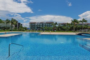 A swimming pool with a clear blue water surface, surrounded by palm trees and a building with multiple balconies. - 2BR Beachfront Condo Playa Pool & Beach (Playa del Carmen)