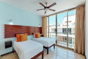 A hotel room with two beds, a ceiling fan, a large window with curtains, and a view of buildings outside. - 2BR Playa Condo: Rooftop Pool, Steps to 5th Ave (Playa del Carmen)
