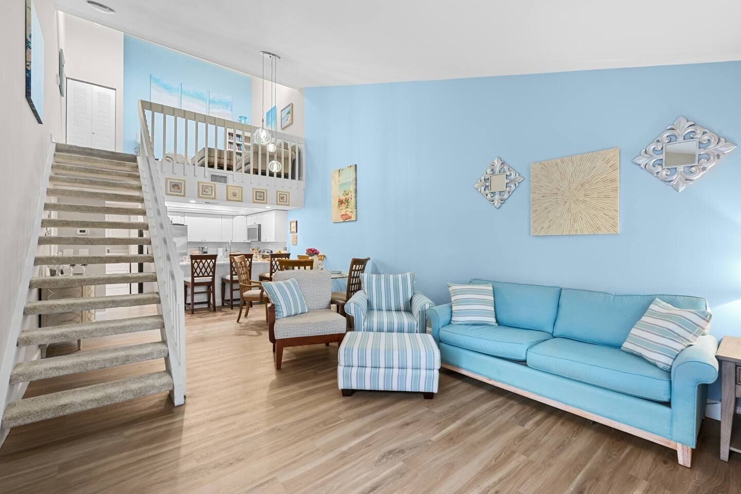 Een ruime woonkamer met een blauwe bank, gestreepte fauteuils, een eettafel en een trap naar een hoger niveau.