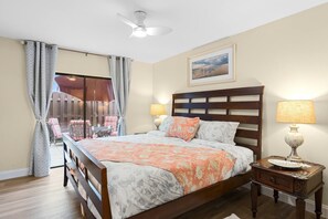 Quarto com cama, mesa de cabeceira, abajur, ventilador de teto e janela com cortinas.