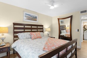 A bedroom with a wooden bed, two bedside lamps, a ceiling fan, a framed landscape painting, and a mirror. - Coastal Glam / Near Beach & Downtown / Heated Pool (Fort Myers)