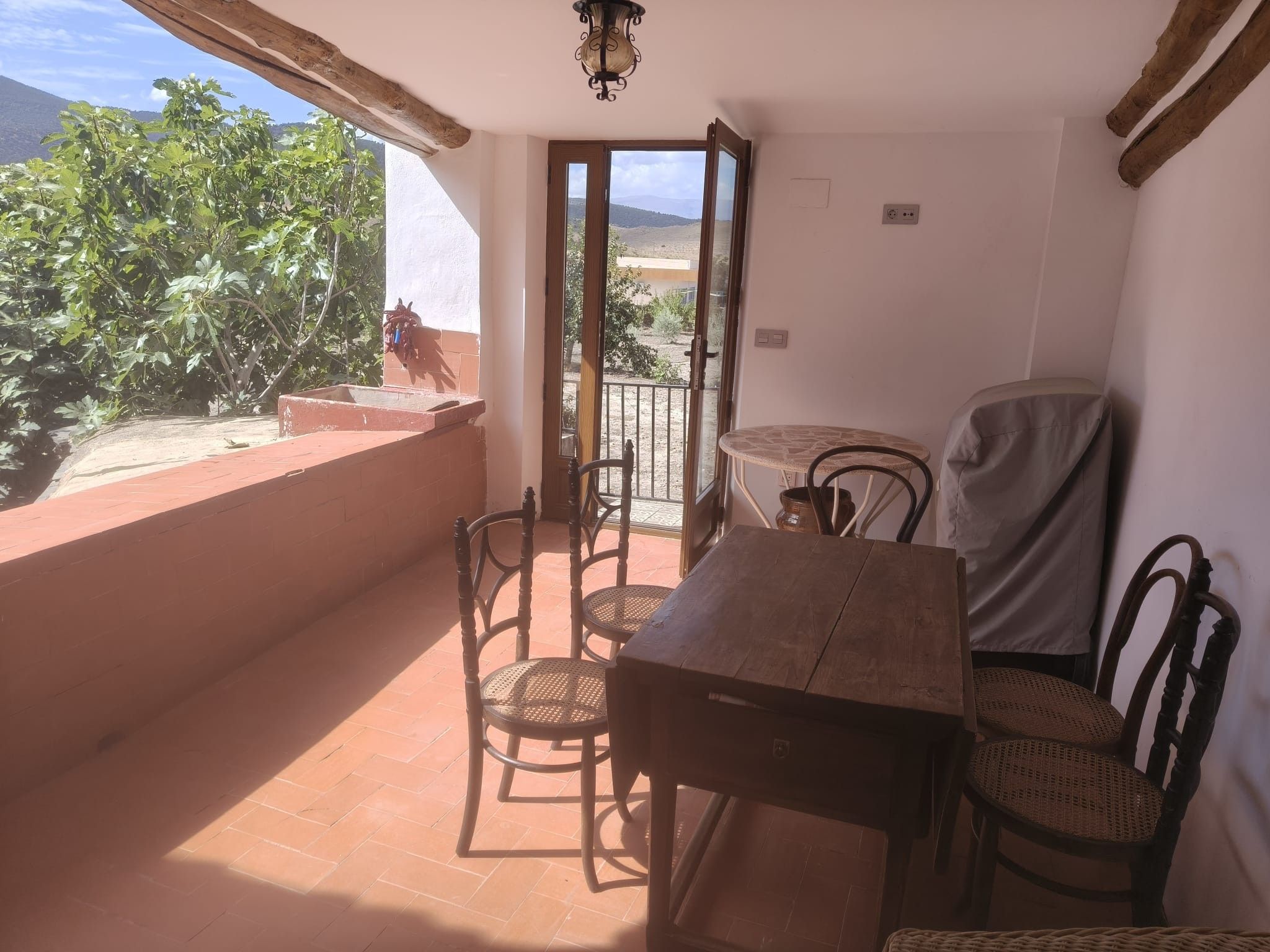 Een balkon met een tafel en stoelen, uitzicht op groen en bergen, en een deur naar buiten.