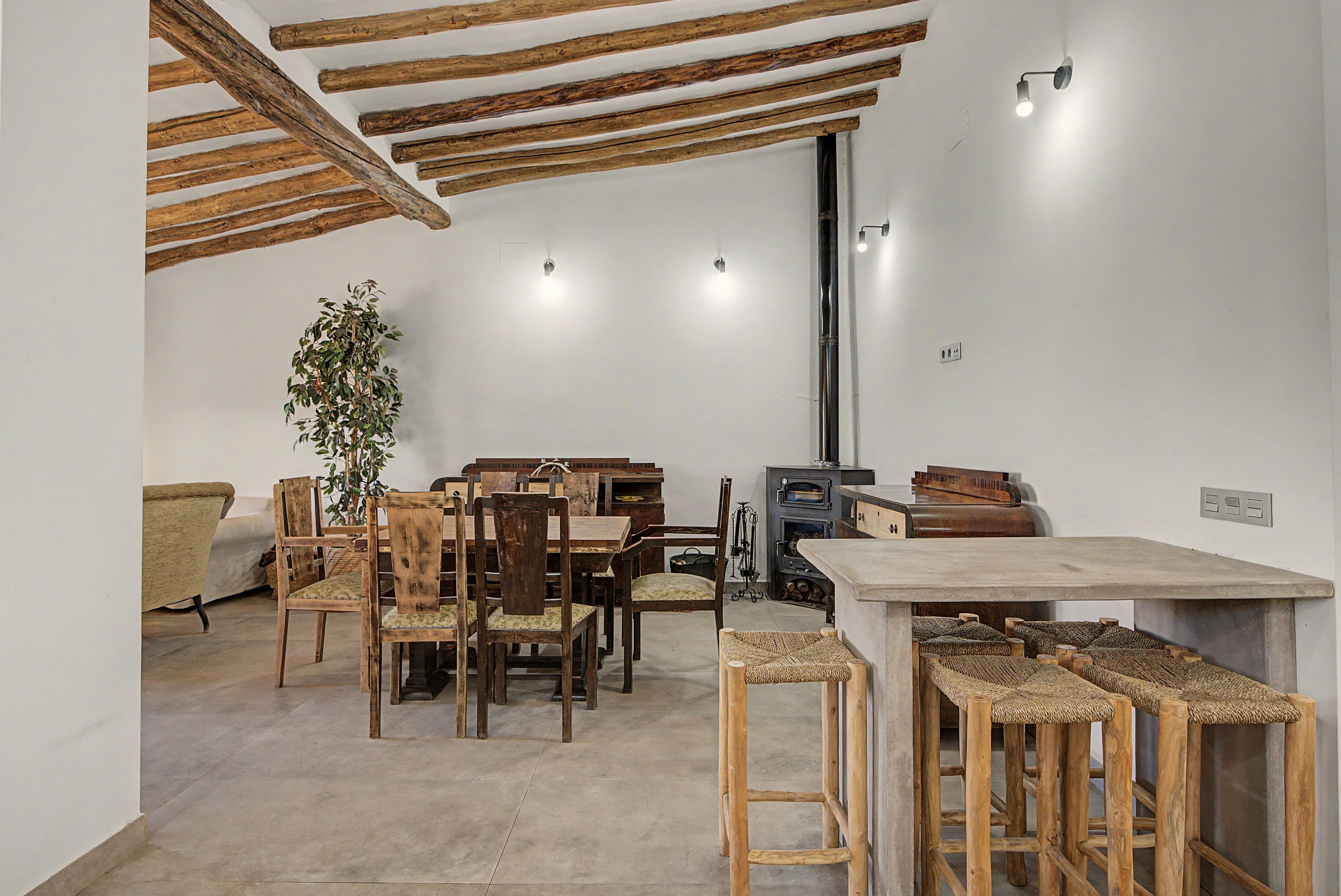 Un intérieur rustique avec des poutres apparentes, un coin repas avec des chaises et des tables en bois, un poêle à bois encastré et une plante en pot.