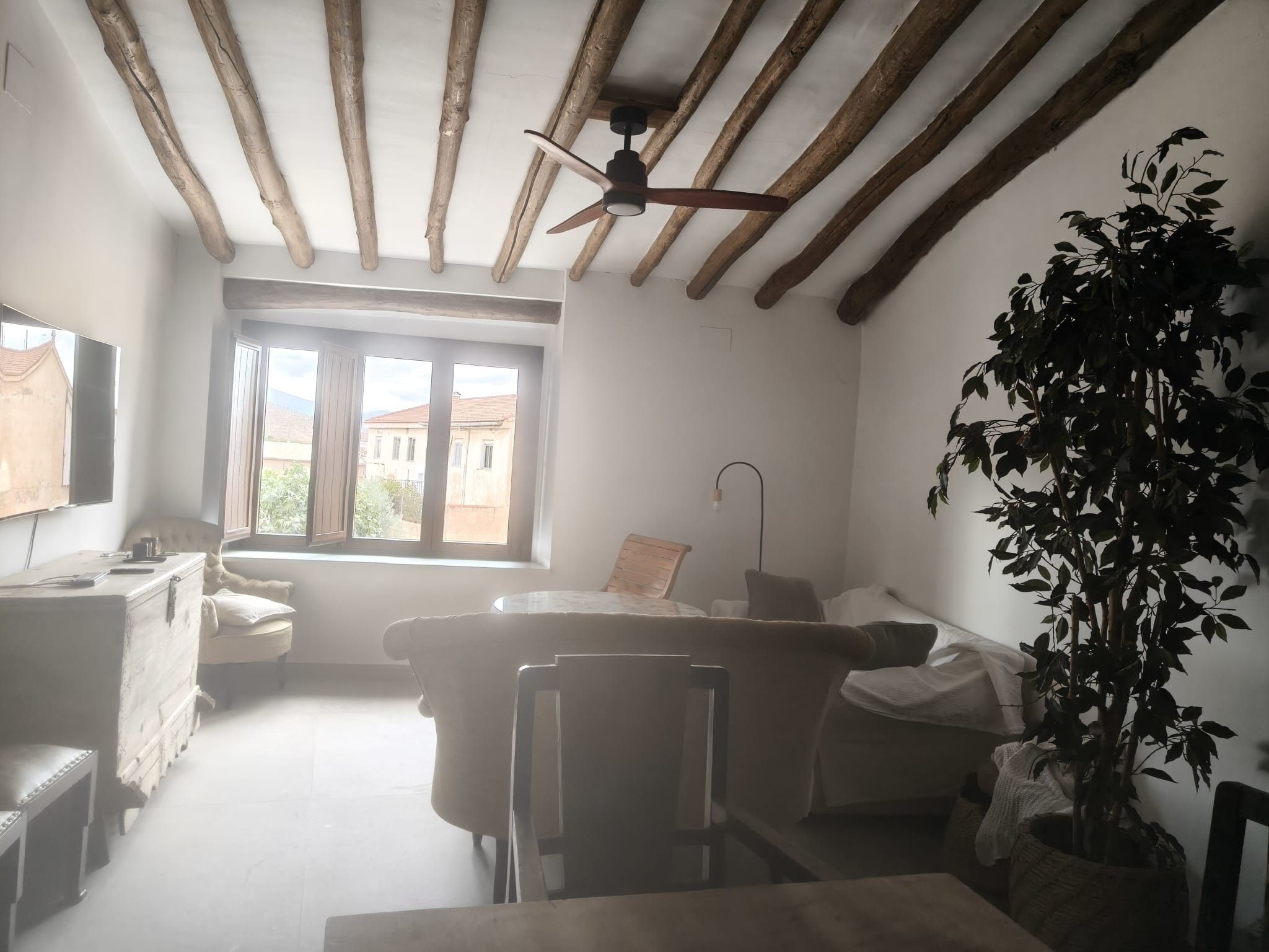 A room with a ceiling fan, a window with a view of buildings, a plant, and a table with chairs.