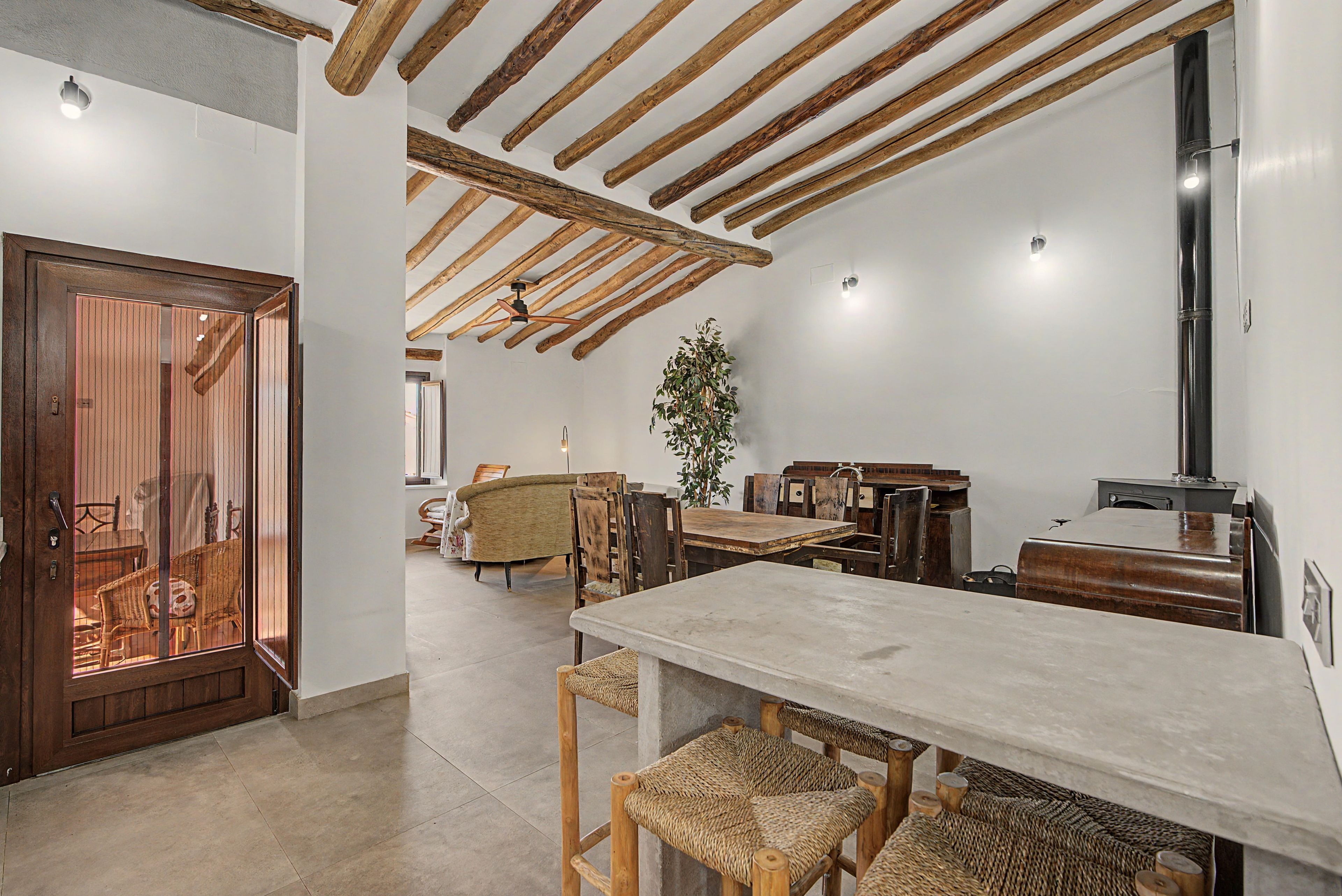 A spacious room with a high ceiling, wooden beams, a dining table, wicker chairs, and a large potted plant.