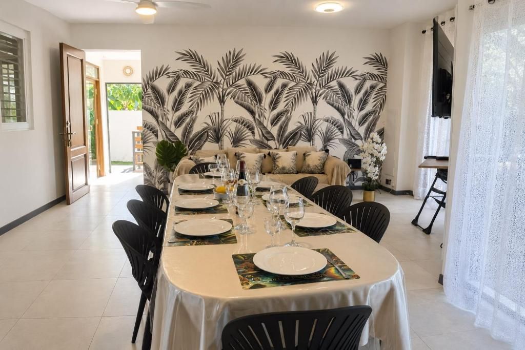 Un espace repas avec une longue table dressée pour un repas, des chaises et un mur décoratif orné de motifs de feuilles de palmier.
