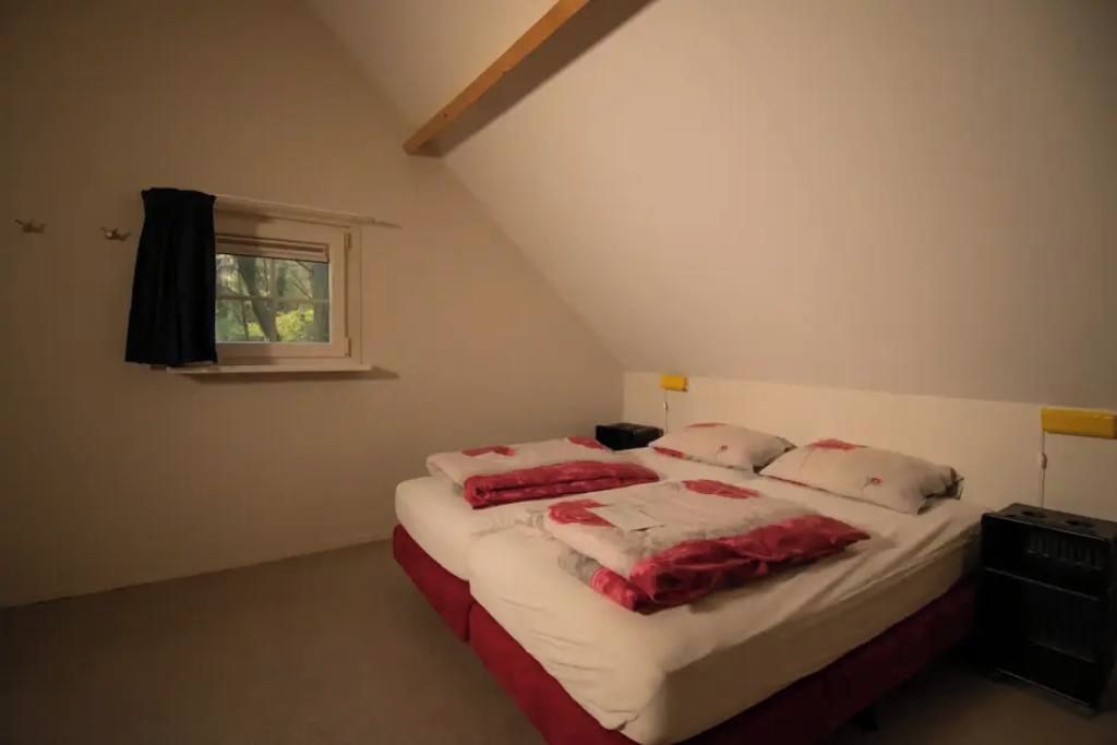 A bedroom with a sloped ceiling, a single bed with white and red bedding, a nightstand, a window with a black curtain, and a small shelf above the bed.