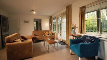 A living room with a sofa, armchair, and coffee table. There is a window with curtains and a view of greenery outside.