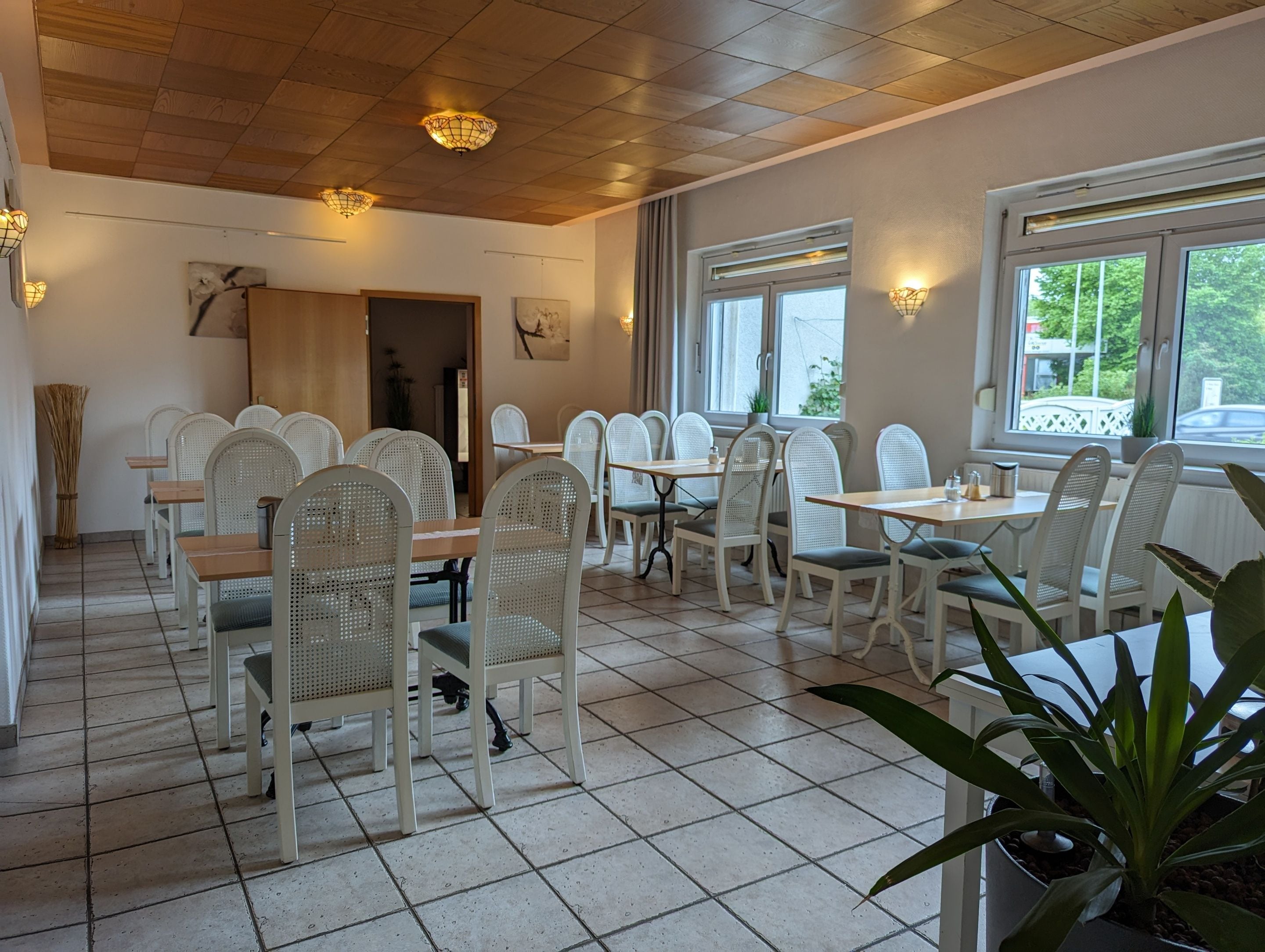 Ein Restaurant mit weißen Korbstühlen und Tischen, Holzboden und großen Fenstern.